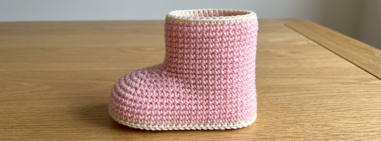 A pink crochet baby boot with a cream sole edge and a cream band round the top, on a wooden table.